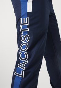 Calças desportivas de tecido azul-marinho com uma risca azul na lateral. Apresenta um grande texto branco "LACOSTE" e um pequeno detalhe branco na parte superior.