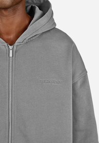 Sweatshirt cinzenta com fecho éclair, feita de material macio, com capuz grande e logótipo "PEGADOR" bordado no peito. Textura suave.