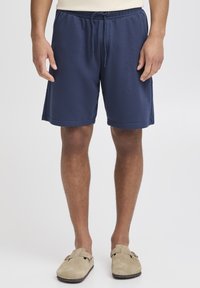 Tamsiai mėlyni sportiniai šortai su elastinga liemene ir raišteliais. Lygios tekstūros su siuvinėtomis apačiomis, derinami su šviesiai rudos spalvos slip-on tipo batais.