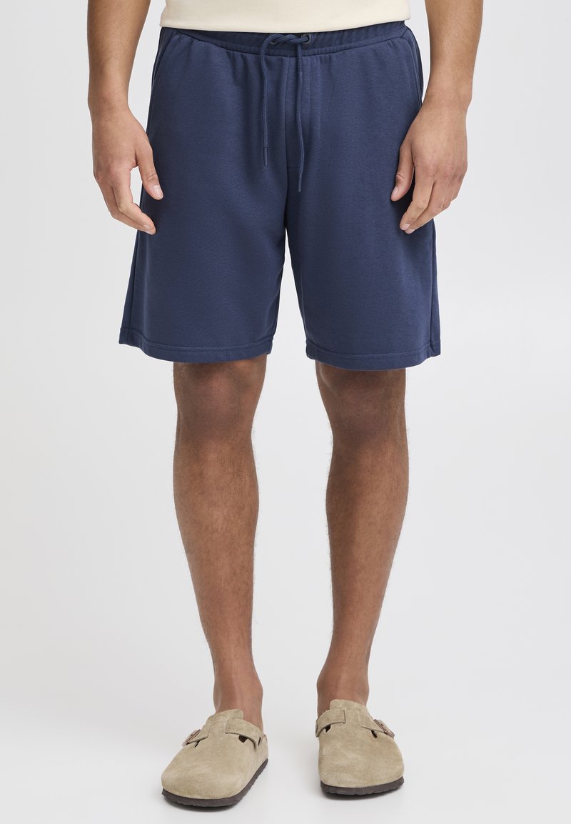 Tamsiai mėlyni sportiniai šortai su elastinga liemene ir raišteliais. Lygios tekstūros su siuvinėtomis apačiomis, derinami su šviesiai rudos spalvos slip-on tipo batais.