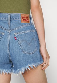 Bermudas de mezclilla con dobladillo deshilachado, de color azul claro, y una etiqueta de cuero con el logo de Levi's en la parte trasera de la cinturilla. Dos bolsillos traseros visibles.