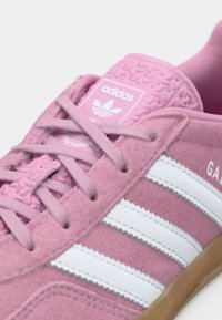 Lys rosa ruskindssneakers med hvide striber, tekstureret gummidetalje på hælen og lyserøde snørebånd. Har et Adidas-logo på tungen.