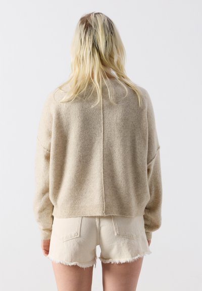 Femme blonde portant un pull beige oversize et un short en denim écru effiloché, debout de dos sur un fond blanc.