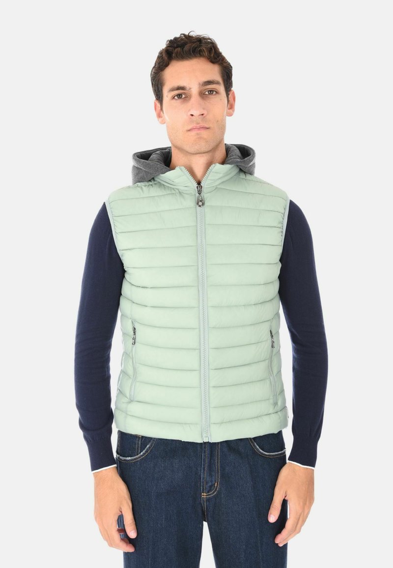 Uomo che indossa un gilet trapuntato verde chiaro sopra una felpa con cappuccio blu navy e jeans, in piedi contro uno sfondo bianco semplice, rivolto in avanti.