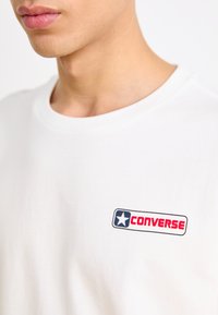 Camiseta blanca de algodón con un logo rectangular que presenta una estrella azul y la palabra "CONVERSE" en letras rojas y negras.