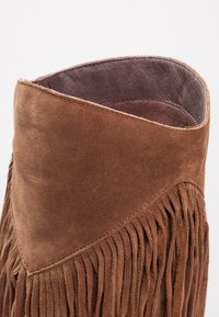 Botín de ante marrón con un diseño de flecos, una parte superior ligeramente redondeada y un suave forro interior gris. Textura suave, forma cónica y acentos mínimos.