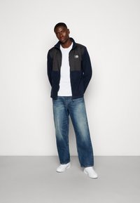 Veste zippée bleu marine et gris avec un col haut, assortie à un t-shirt blanc et un jean large bleu clair, portée avec des baskets blanches.