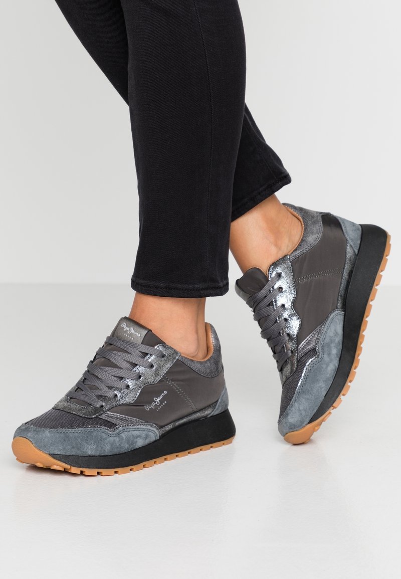 Personne portant un pantalon noir slim et des baskets grises avec des détails en daim, mesh et métal, ainsi que des semelles en caoutchouc naturel, debout sur un sol clair.