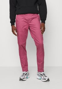 Mężczyzna w różowych spodniach, czarnej bluzie i białych butach sportowych stoi na tle jasnego, gładkiego tła.