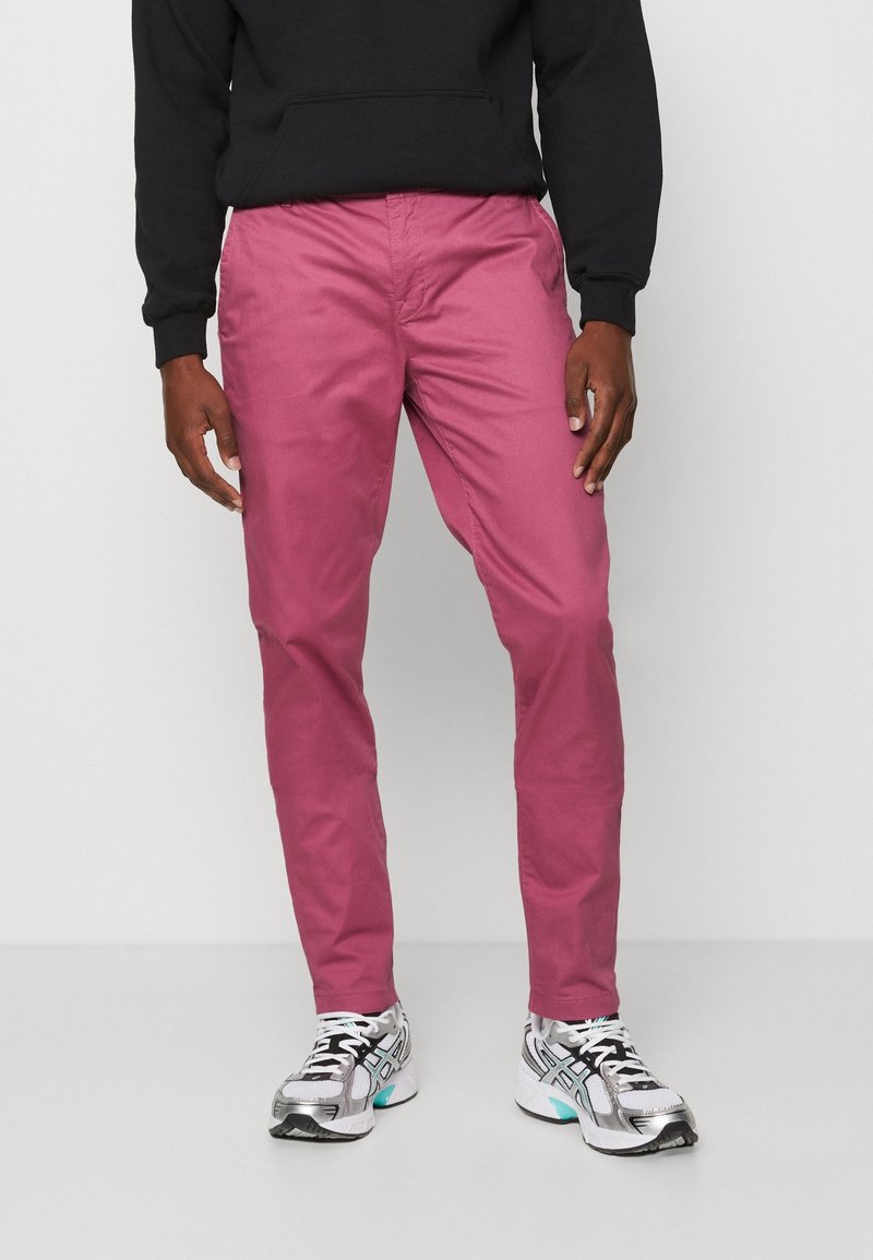 Mężczyzna w różowych spodniach, czarnej bluzie i białych butach sportowych stoi na tle jasnego, gładkiego tła.