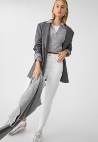 Šedý blazer, světle šedý zkrácený svetr, bílé džíny a stříbrné tenisky; drží šedou pletenou módní kousek. Hladké textury a striktní design.
