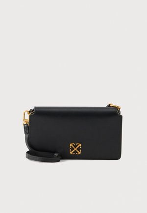 Bolso de hombro de piel negra con solapa, herrajes en tono dorado y un emblema dorado de flechas cruzadas, sobre un fondo claro.