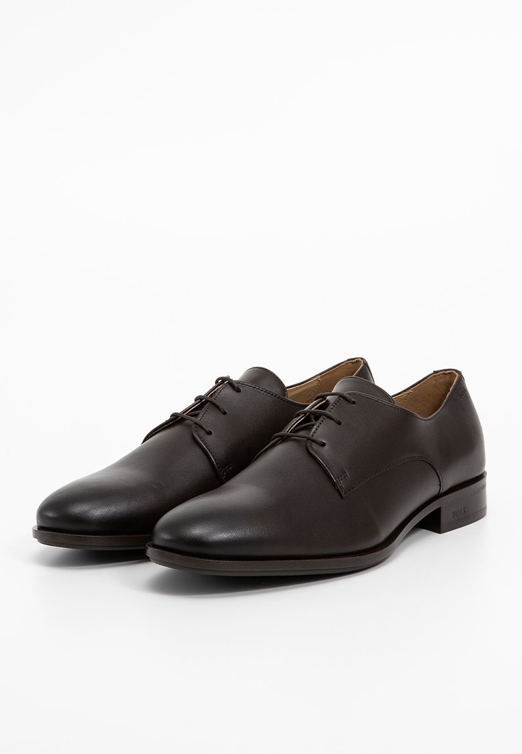 Des chaussures de costume en cuir marron présentent une finition lisse, un bout arrondi, un design à lacets et un talon bas. Construction durable avec des coutures discrètes.