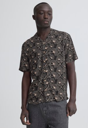 Hombre con una camisa negra de manga corta con botones, con estampado floral beige y vaqueros gris oscuro, mirando hacia abajo.