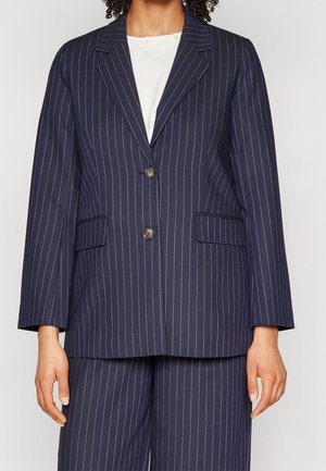 Kvinde iført en navyblå jakkesæt med smalle striber, to-knaps blazer og matchende vidde bukser over en hvid bluse.