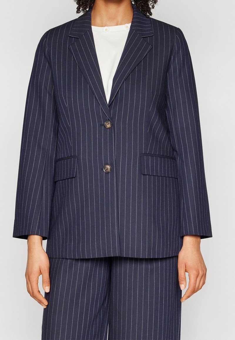 Femme portant un costume bleu marine à fines rayures, composé d'un blazer à deux boutons et d'un pantalon à jambes larges assorti, avec un chemisier blanc.