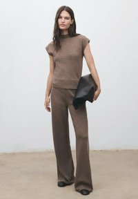 Top lavorato a maglia marrone con maniche a cappuccio e pantaloni larga abbinati. La modella tiene una pochette nera. Trama liscia, design semplice.