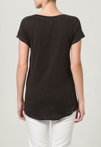 Woman wearing a black short-sleeve t-shirt with white stitching and white pants, shown from the back against a neutral background.