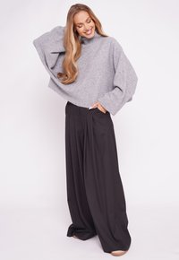 Szary, oversize sweter z wysokim kołnierzem, z prążkowanymi mankietami, oraz czarne, szerokie spodnie z zakładkami, na jednolitym białym tle.