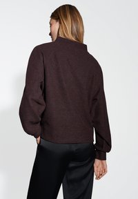 Giacca cropped in misto lana marrone con collo alto, maniche raglan e polsini a costine. Indossata sopra pantaloni neri in satin.