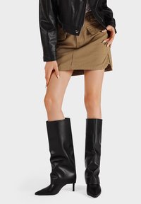 Botas de cuero negras hasta la rodilla con un diseño arrugado, punteras puntiagudas y delgados tacones de aguja. Llevadas con una falda caqui y una chaqueta negra.