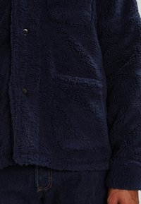 Granatowa polarowa kurtka z guzikami z przodu i kwadratową kieszenią, noszona z ciemnoniebieskimi dżinsami.