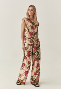 Vrouw in een beige twee-delig outfit met grote rode en groene bloemmotieven, staand met ontspannen handen, kijkend opzij, tegen een effen lichte achtergrond.