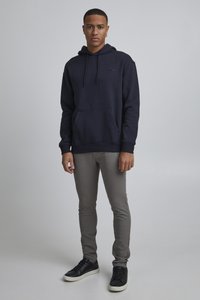 Ung mand står op, iført en mørkeblå hoodie, grå slim-fit bukser og sorte sneakers med hvide såler, mod en ensfarvet grå baggrund.