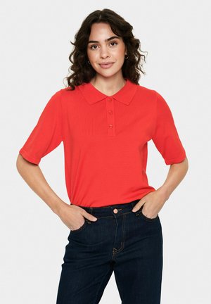 T-shirt polo rouge à manches courtes, col classique, patte de boutonnage à trois boutons et texture lisse, associé à un jean bleu marine.