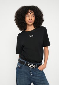 Femeie cu păr creț purtând un tricou negru cu un logo, asortat cu blugi albaștri și o curea neagră cu o cataramă argintie.