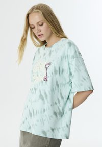 Camiseta de tie-dye en azul claro con mangas cortas, que presenta un gráfico de llave y texto, adornada con pequeños brillantitos para añadir detalle.