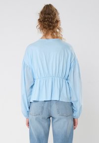 Svetlo modra bluza z dolgimi napihnjenimi rokavi, zategnjeno pasom in sproščenim peplum robom. Mehka tkanina z gladko teksturo in preprostim dizajnom.