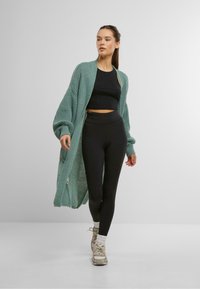 Cardigan long en tricot teal, coupe ample avec des manches oversized, associé à un crop top noir et un legging taille haute. Baskets décontractées.