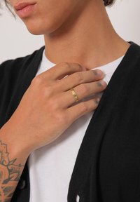 Gold ring on a hand resting on a black cardigan; hand shows tattoo details. The shirt underneath is white.