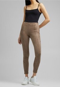 Vrouw in bruin slim-fit suède broek, zwart mouwloos shirt en witte sneakers, staand tegen een effen grijze achtergrond.