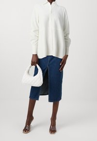 Camicia bianca con bottoni, abbinata a una gonna in denim scuro con spacco frontale. Accentuata con una borsa bianca trapuntata e sandali con tacco trasparente.