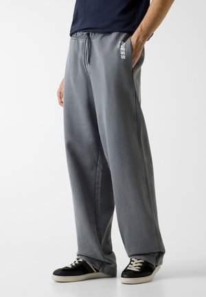 Pantaloni della tuta grigi a vestibilità ampia, con vita regolabile tramite coulisse, tasche laterali e un dettaglio logo bianco. Abbinati a sneakers nere con lacci color crema.