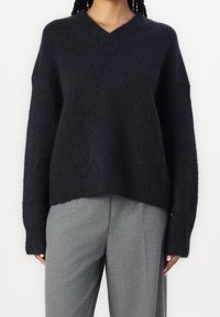 Femme portant un pull en maille duveteuse bleu marine à encolure en V avec manches longues et un pantalon gris ample, debout devant un fond uni.