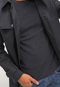 Svart trench coat med knapplås, bärs över en mörkgrå åtsittande skjorta. Synlig hand på kappan som visar en slät tygtextur.