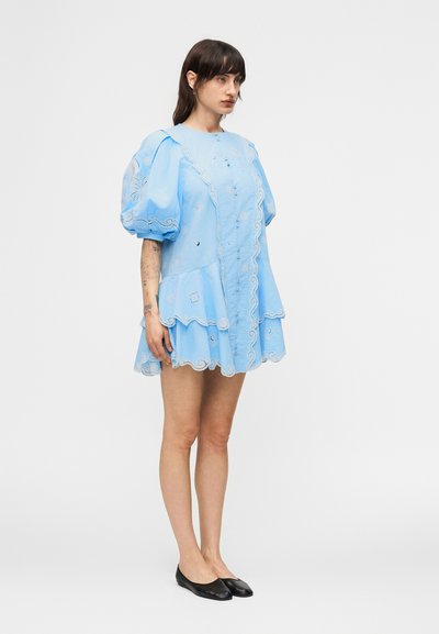 Γυναίκα που φοράει ανοιχτό μπλε κεντητό φόρεμα με φουσκωτά μανίκια και κυματιστό τελείωμα, φοράει μαύρα επίπεδα παπούτσια, στέκεται μπροστά σε λευκό φόντο.