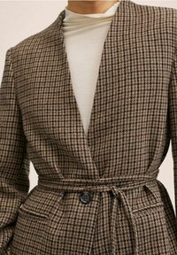 Blazer marrón a cuadros con un ajuste entallado, cintura ceñida y cierre de un solo botón. Presenta una textura suave y cuello con solapas.