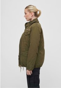 Chaqueta de estilo militar en color verde oliva con cuello alto, cremallera frontal y múltiples bolsillos; cuenta con un cordón en la cintura y una tela texturizada.