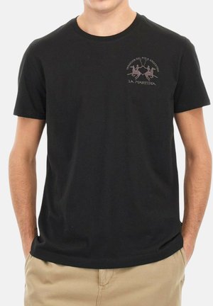 Mand iført sort kortærmet T-shirt med lille polo-spiller logo og tekst på venstre bryst, parret med beige bukser.