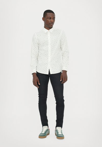 Chemise blanche avec des pois multicolores, col boutonné, coupe slim. Portée avec un jean skinny noir et des baskets vert-bleu avec des lacets blancs.