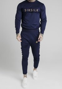 Man i marinblå SikSilk träningsoverall med logotyp på bröstet och benet, i kombination med vita sneakers, stående mot en neutral grå bakgrund.