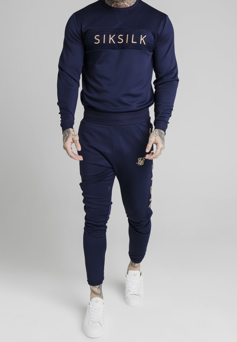 Man i marinblå SikSilk träningsoverall med logotyp på bröstet och benet, i kombination med vita sneakers, stående mot en neutral grå bakgrund.