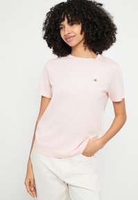 Camiseta de algodón color rosa claro con mangas cortas, cuello redondo y un pequeño logo bordado en el pecho izquierdo. Textura suave, ajuste relajado.