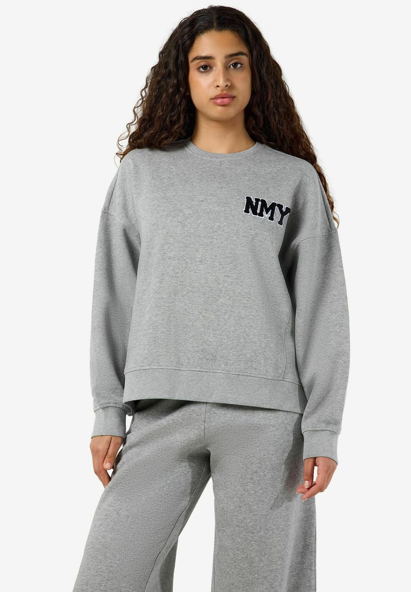 Jeune femme aux longs cheveux bouclés portant un sweat-shirt gris avec "NMY" en lettres noires et un pantalon de survêtement gris assorti.
