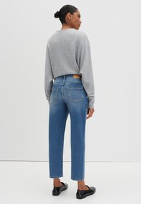 Femme de dos, portant un haut à manches longues gris clair, un jean taille haute bleu et des chaussures noires à enfiler, sur fond blanc.