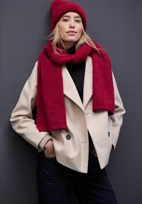 Abrigo de lana crema cruzado con doble botonadura sobre un suéter de cuello alto negro, combinado con una bufanda texturizada roja y un gorro a juego, sobre un fondo gris.